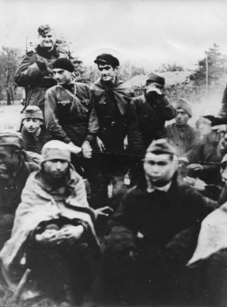 Obóz jeniecki w Szebniach - Grupa jeńców 1941-42 grzejących się przy zapalonym ognisku. Niemiecki wartownik uzbrojony w karabin w płaszczu i furażerce.